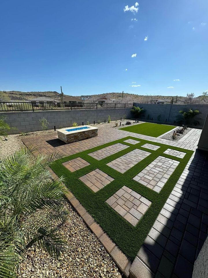A backyard with a geometric pattern of brick pavers and artificial grass showcases expert paver installation Phoenix AZ, featuring a rectangular fire pit, desert landscaping, and distant hills under a clear blue sky.