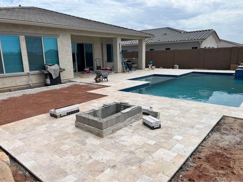 A backyard with a swimming pool and light stone tile patio under construction, showcasing Paver Installation Phoenix AZ, features a partially built rectangular structure and building materials scattered around. A covered patio and house are in the background.
