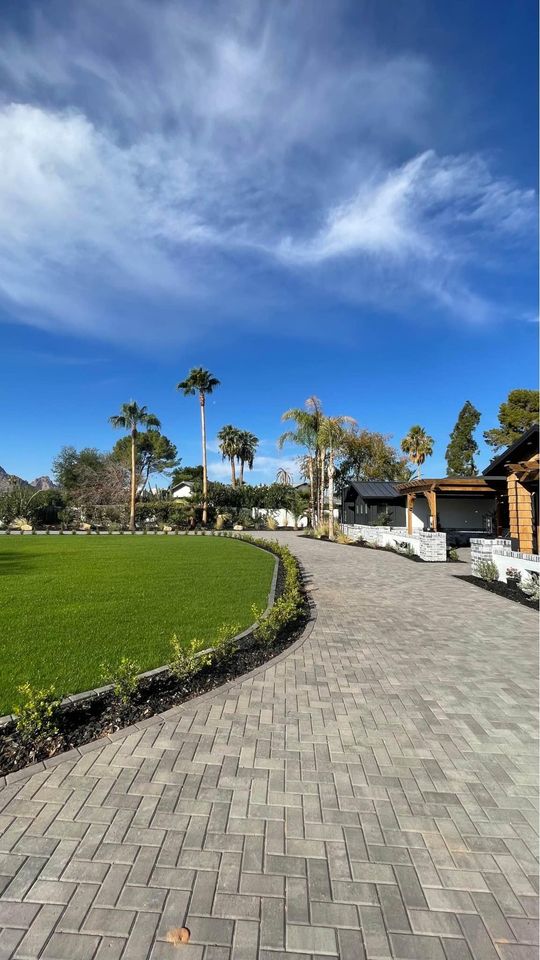 A paved walkway, crafted with expert paver installation Phoenix AZ, curves alongside a green lawn dotted with palm trees under a blue sky. Modern buildings rise to the right, while landscaped shrubs neatly border the path.