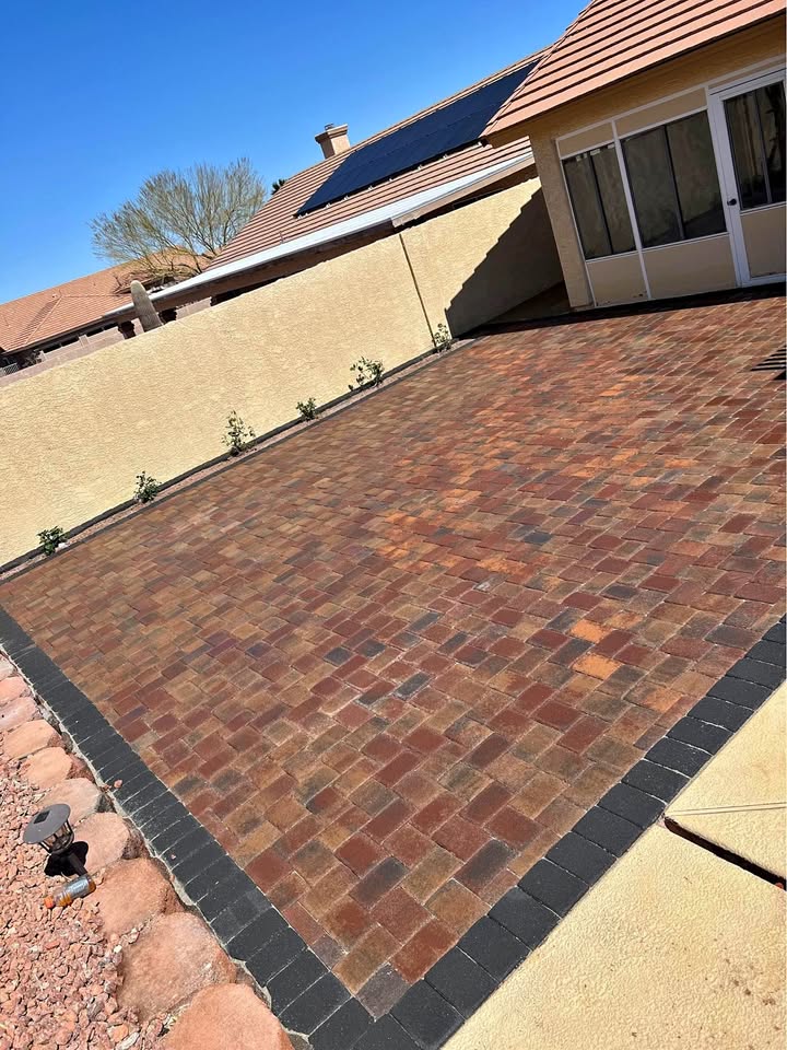 A backyard with a patio made of red and brown rectangular pavers, bordered by black pavers—showcasing expert Paver Installation Phoenix AZ—next to a tan house with solar panels and a beige wall lined with small plants.