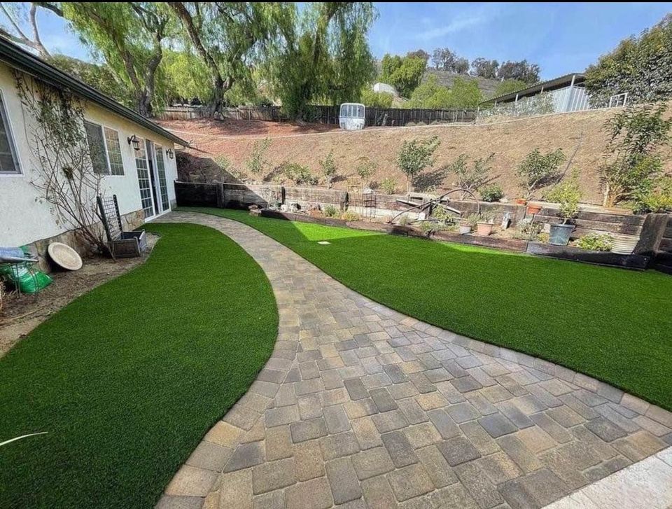 A stone walkway, created with expert paver installation Phoenix AZ, curves through a backyard with artificial green grass and garden beds along a sloped hill behind a white house. Trees and shrubs grow near the fence at the top of the slope.