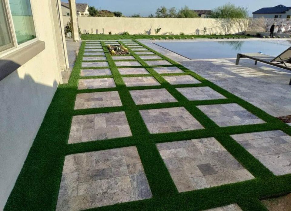 A modern patio features large square stone tiles set in a grid pattern with green grass borders, next to a swimming pool, lounge chair, and a light-colored house.