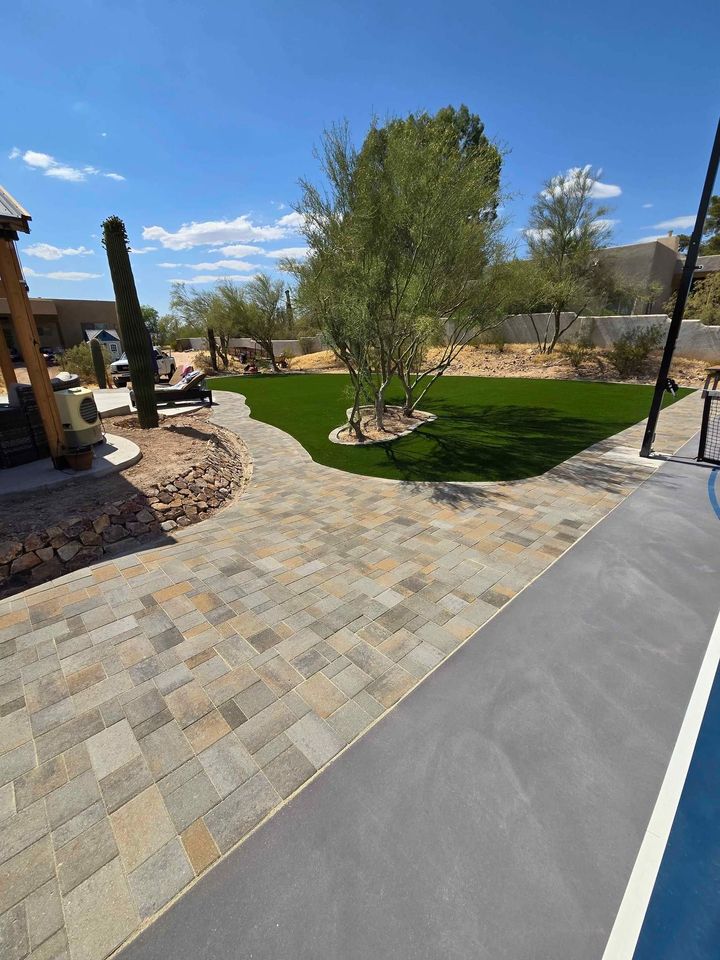 A sunny backyard with a stone patio, artificial grass lawn, desert landscaping, trees, cacti, and a pool edge in the foreground, surrounded by a low wall and a clear blue sky above.