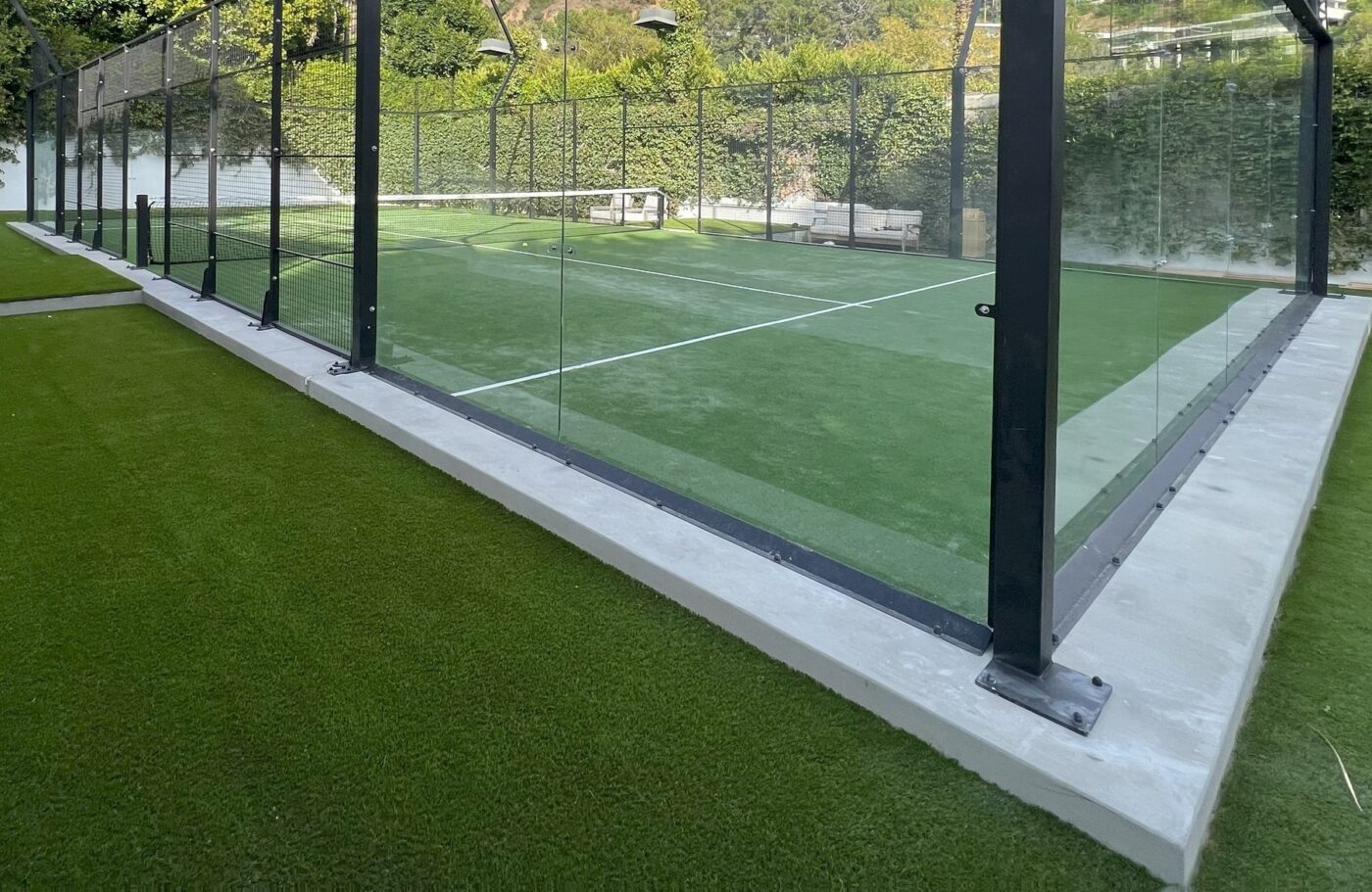 A padel tennis court with glass walls and green artificial turf is surrounded by lush greenery and enclosed by a tall fence under outdoor lighting.