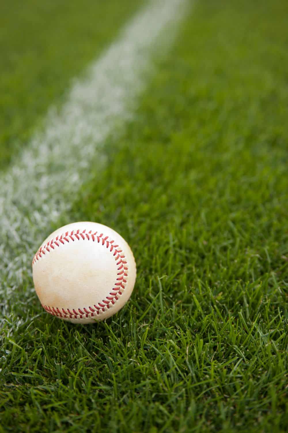 A white baseball with red stitching rests on green grass near a white foul line on a baseball field.