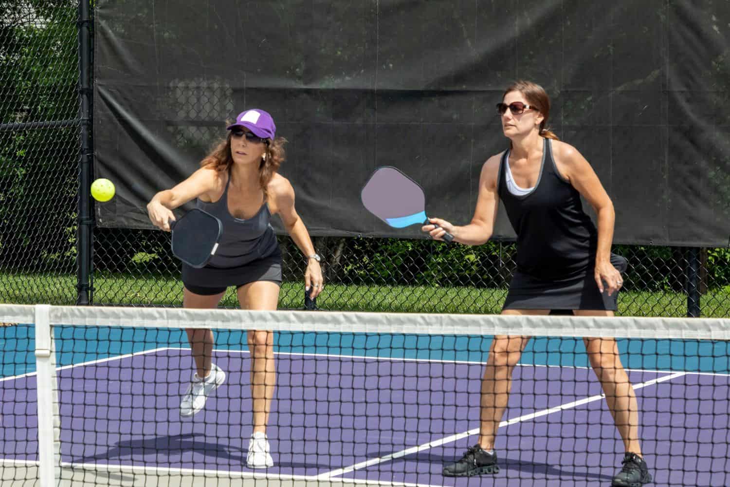 Two women playing pickleball on an outdoor court, both holding paddles and watching a ball mid-air. One woman is lunging forward to hit the ball, while the other stands ready behind her.