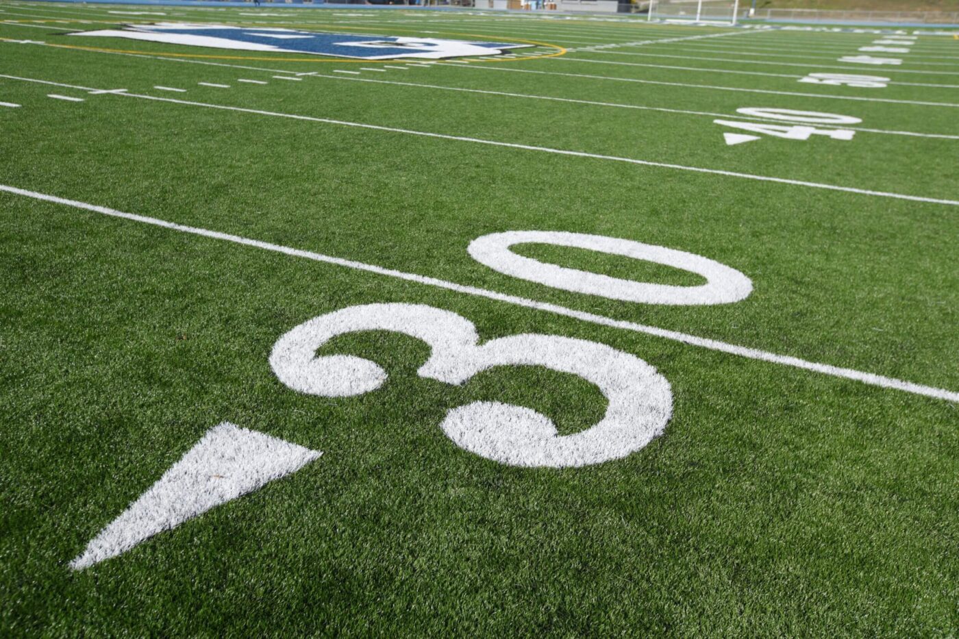 Close-up view of the 30-yard line on a green football field, with white boundary lines and part of a blue and white midfield logo visible in the background.
