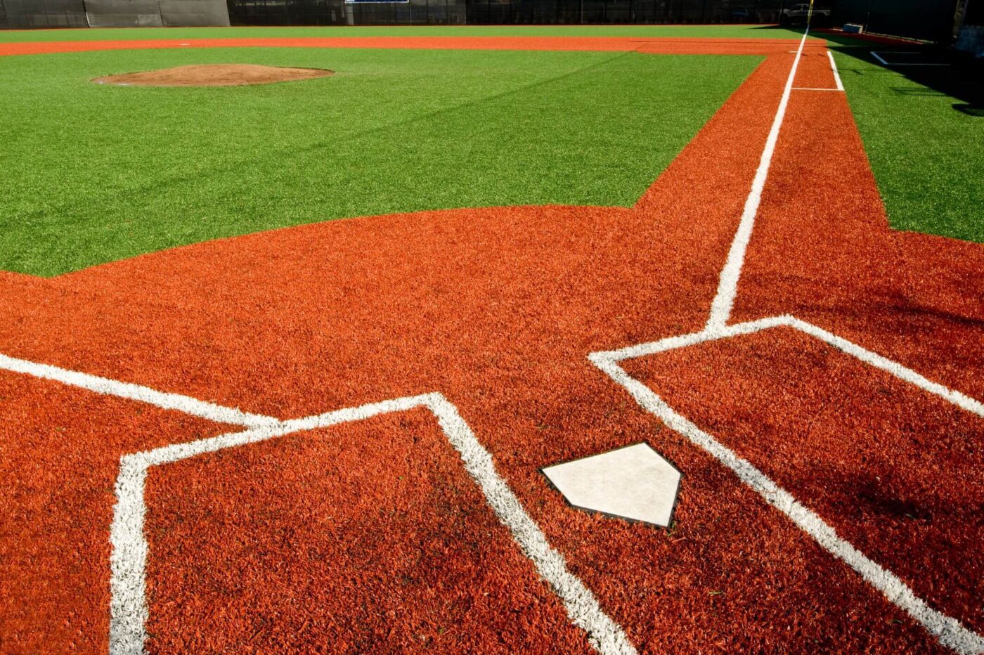Close-up view of home plate and the batter's box on a baseball field with bright red artificial turf and clear white lines; green turf outfield and pitcher's mound visible in the background.