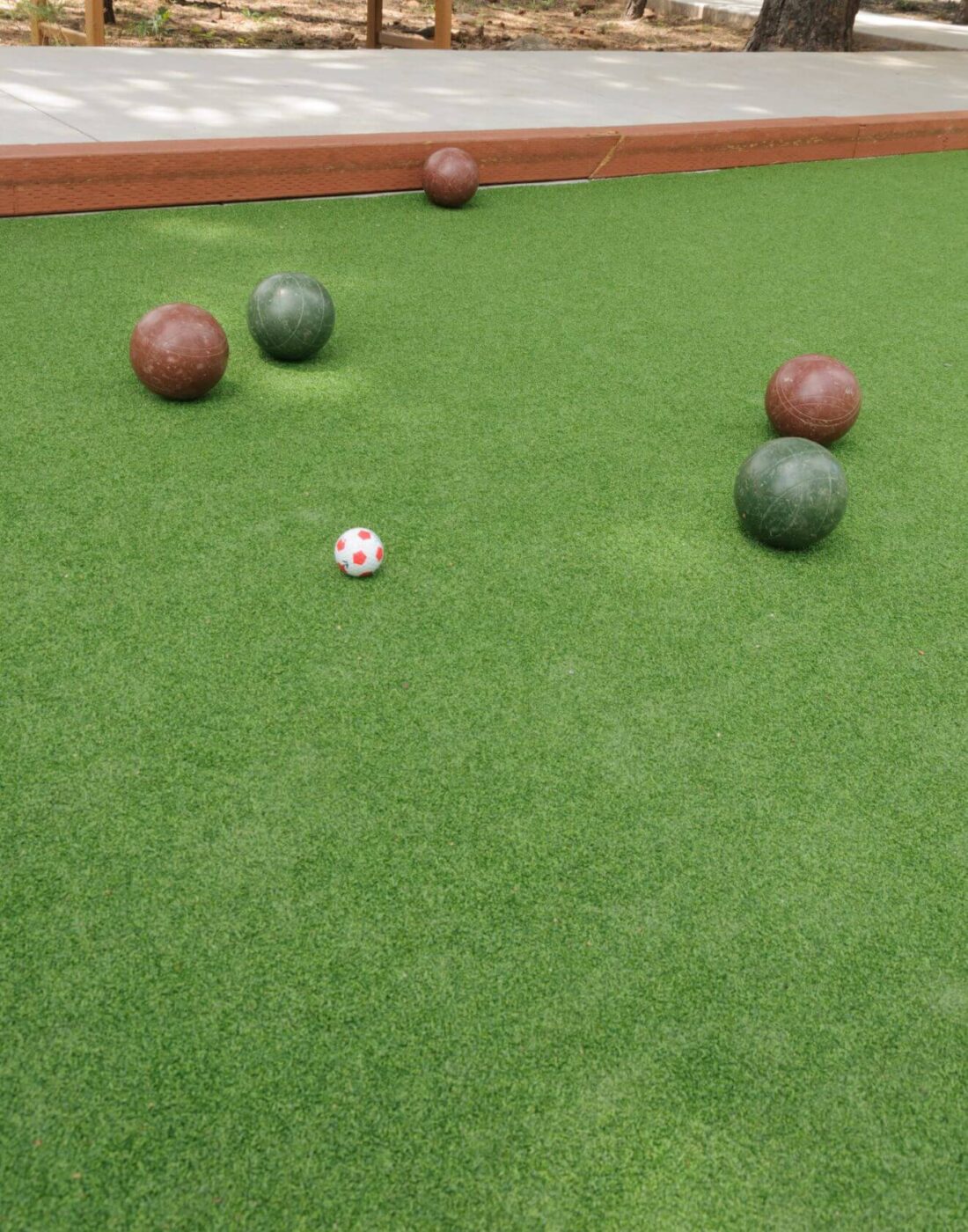 Four bocce balls—two green and two brown—are scattered on a green artificial turf bocce court. A small white and red pallino ball is near the center of the image. A wooden border lines the court.