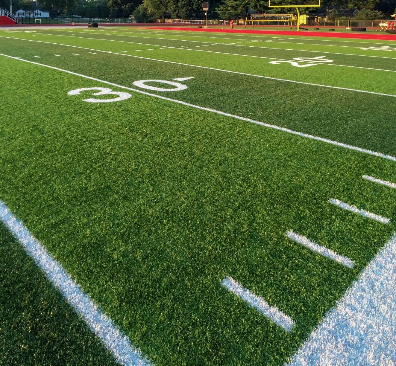 A view of a football field with green artificial turf, white yard lines, and numbers marking the 30-yard line. The yellow goalpost and red running track are visible in the background.