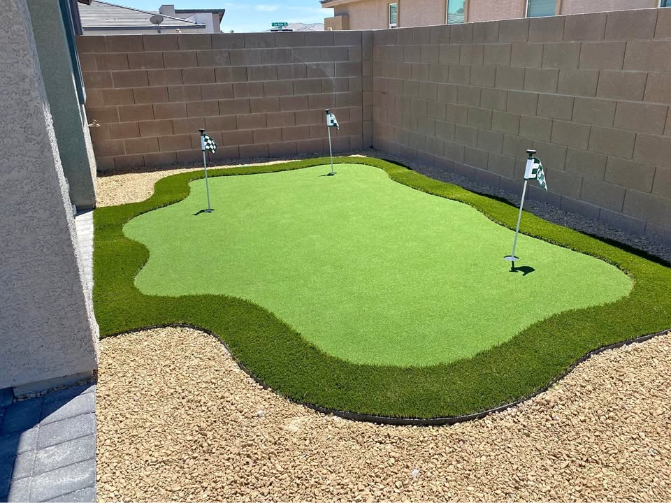 A small backyard putting green with three golf holes and flags, surrounded by artificial turf and gravel, enclosed by tan brick walls and neighboring houses.