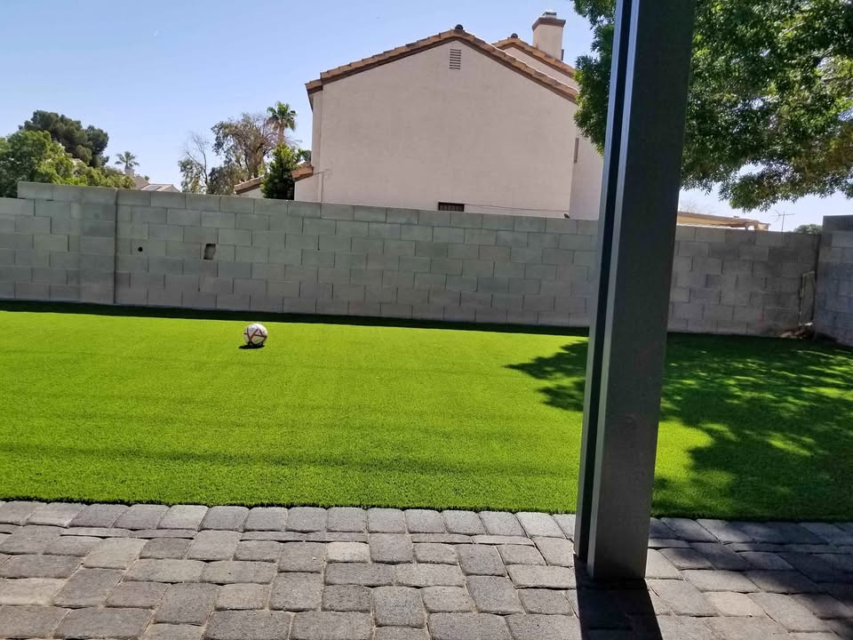 A backyard with bright green artificial grass, a single soccer ball on the lawn, a gray brick patio in the foreground, and a tall cinder block wall enclosing the yard. A house and trees are visible beyond the wall.