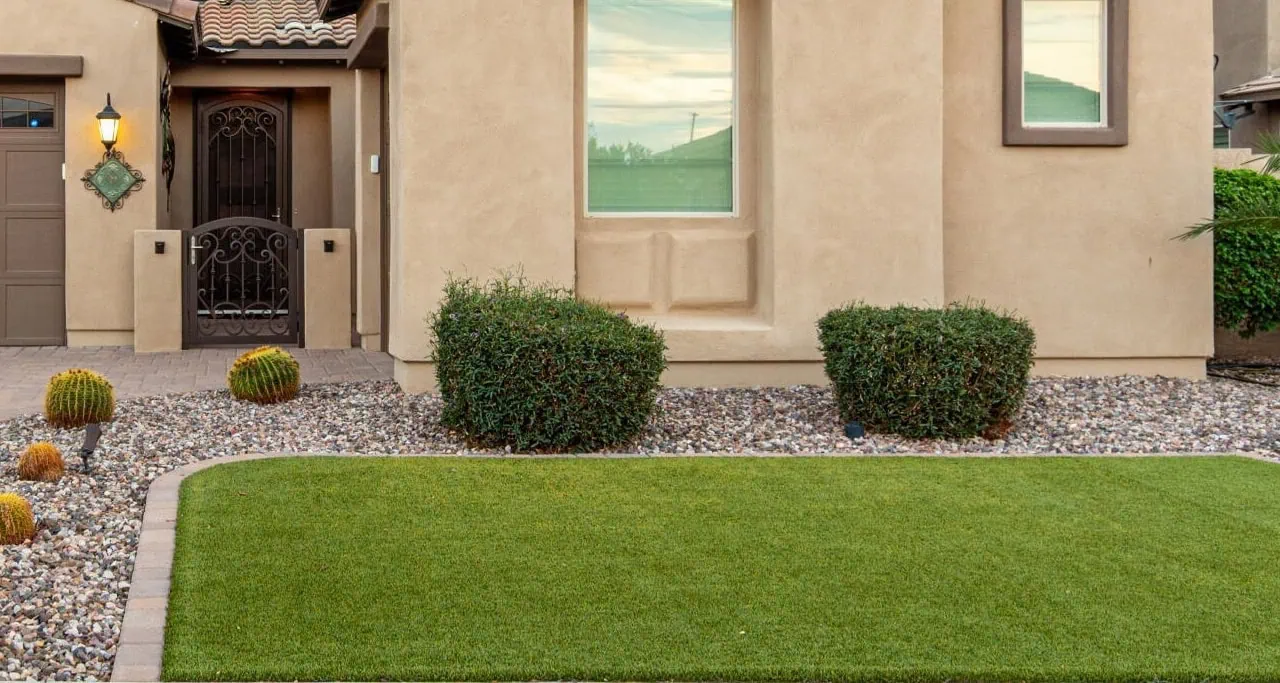 A neatly landscaped front yard features green artificial grass, decorative rocks, rounded bushes, small cacti, and the tan exterior of a modern house with a window and a wrought-iron door.