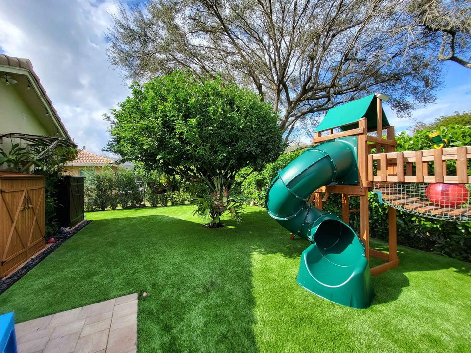 A backyard playground with artificial grass in Phoenix, AZ, features a large leafy tree and a wooden playset with a green spiral tube slide and small bridge, surrounded by lush plants, a wooden fence, and a house on the left.