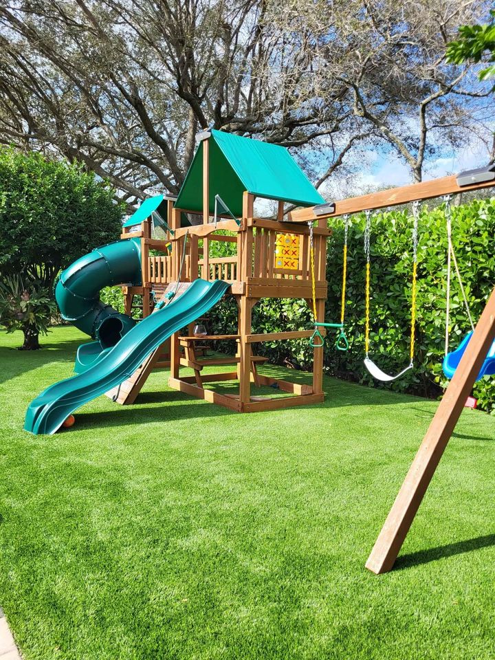 A wooden backyard playset with green slides, swings, rope ladder, and a small fort sits on bright Playground Artificial Grass Phoenix AZ, surrounded by bushes and trees under a blue sky.