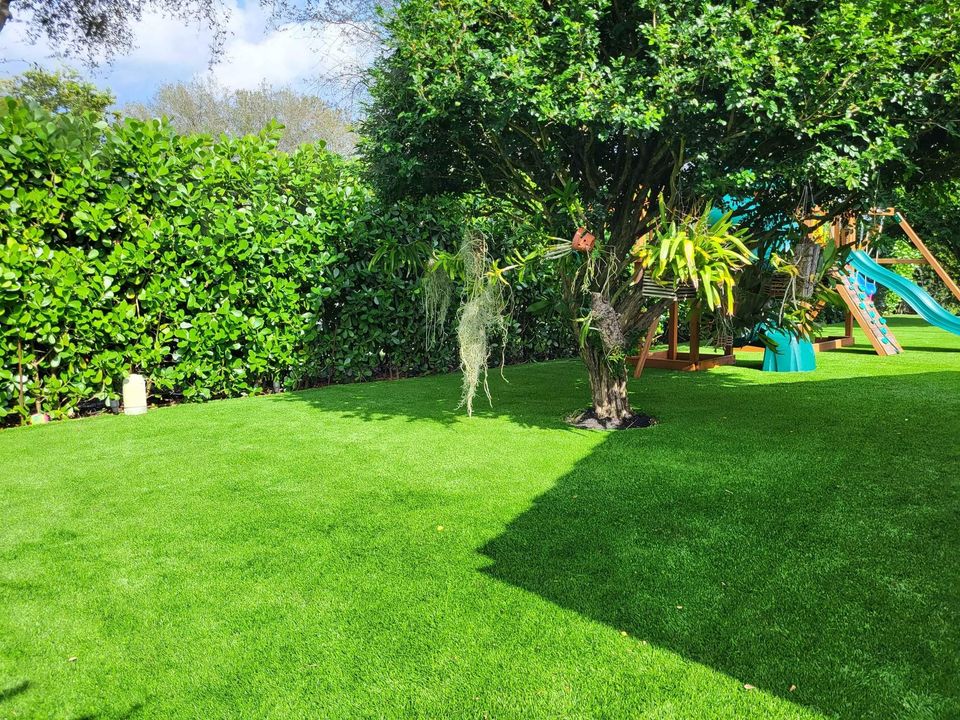 A sunny backyard with Playground Artificial Grass Phoenix AZ, a large tree draped in hanging moss, dense green hedges, and a colorful children’s playset with slides in the background.