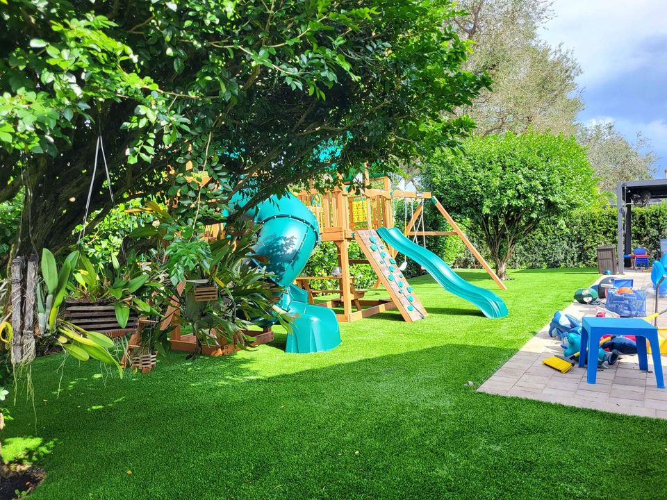 A backyard with Playground Artificial Grass Phoenix AZ, a leafy tree, and a wooden playset featuring a blue slide and climbing elements. Nearby are blue and red plastic chairs and children's toys scattered on a paved area.