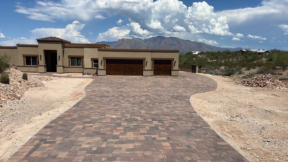 A modern, single-story house with a wide paved driveway, surrounded by a desert landscape and distant mountains under a partly cloudy sky.